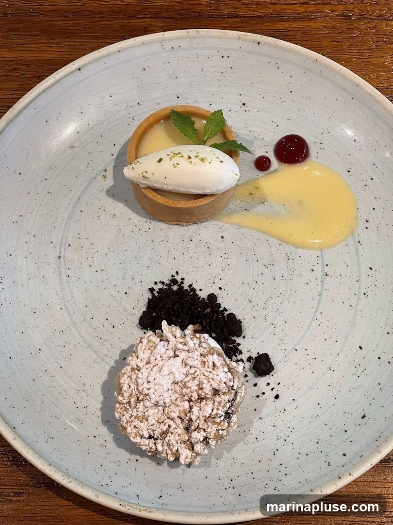 A dessert is presented on a speckled blue plate. The dessert includes a small tart with a dollop of white cream and mint leaves, a swirl of yellow sauce, and a red berry coulis. To the side, there is a mound of crumbled dark cookies and a larger, crispy dessert dusted with powdered sugar.