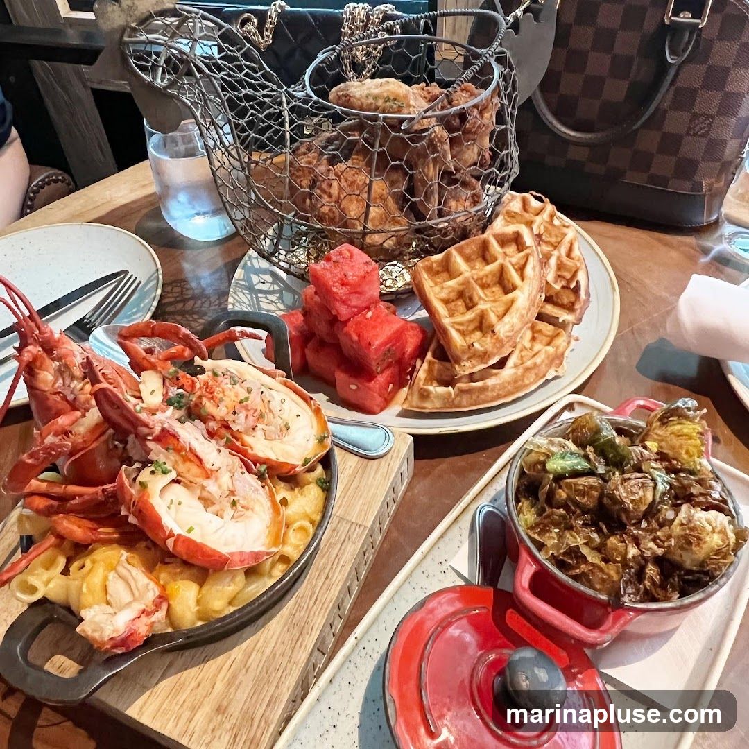 A table laden with a meal featuring lobster mac and cheese, fried chicken in a wire basket, waffles, watermelon cubes, and Brussels sprouts. A handbag is visible in the background.