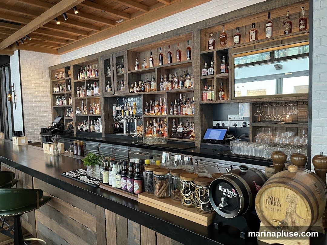 A well-stocked bar interior featuring a long counter and extensive shelving filled with various bottles of liquor. The shelves are made of rustic wood and are organized with different types of spirits. Behind the counter, there are beer taps and a digital ordering system. On the counter, there are small barrels, jars of dried fruit, and bottles of mixers. The ceiling has exposed wooden beams and track lighting.
