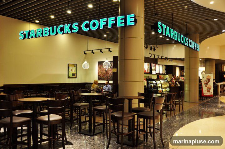 An interior view of a Starbucks coffee shop with prominent signage. The image shows seating areas with tables and chairs, a counter with a display case, and customers. The Starbucks Coffee logo is visible on illuminated signs.