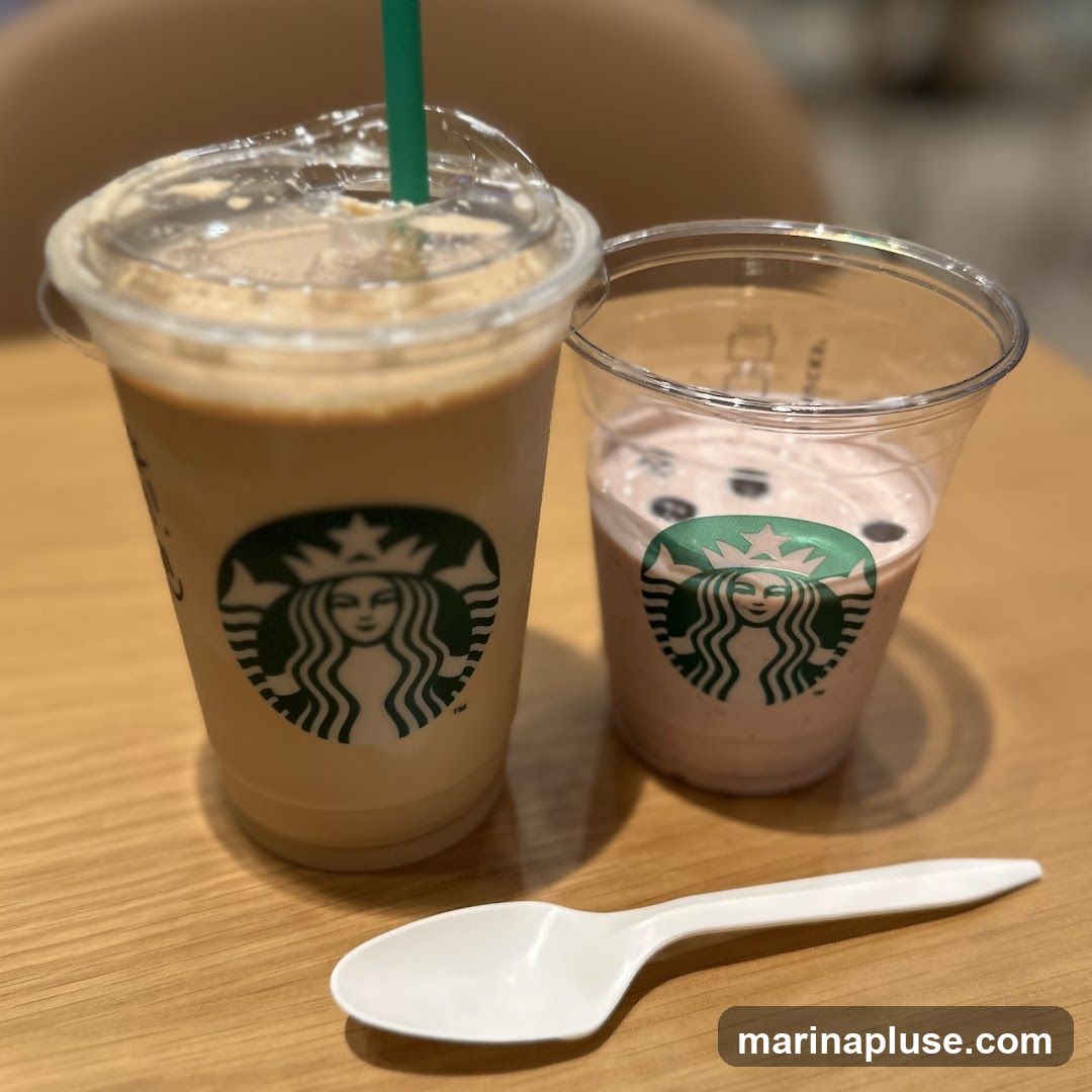 Two Starbucks drinks, one a light brown coffee beverage and the other a pink strawberry-like drink, sit on a wooden table next to a white plastic spoon. Both cups feature the iconic Starbucks siren logo.