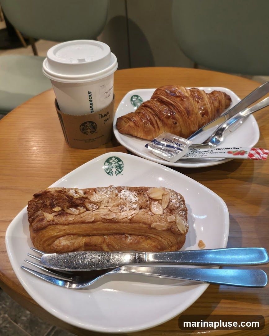 A white paper cup with a lid and a brown cardboard sleeve with the Starbucks logo sits on a wooden table. Next to the cup are two white plates, each with a pastry. The plate in the foreground holds an almond-topped pastry dusted with powdered sugar, with a knife and fork resting on the plate. The plate in the background holds a croissant with a small packet of butter and a packet of jam.