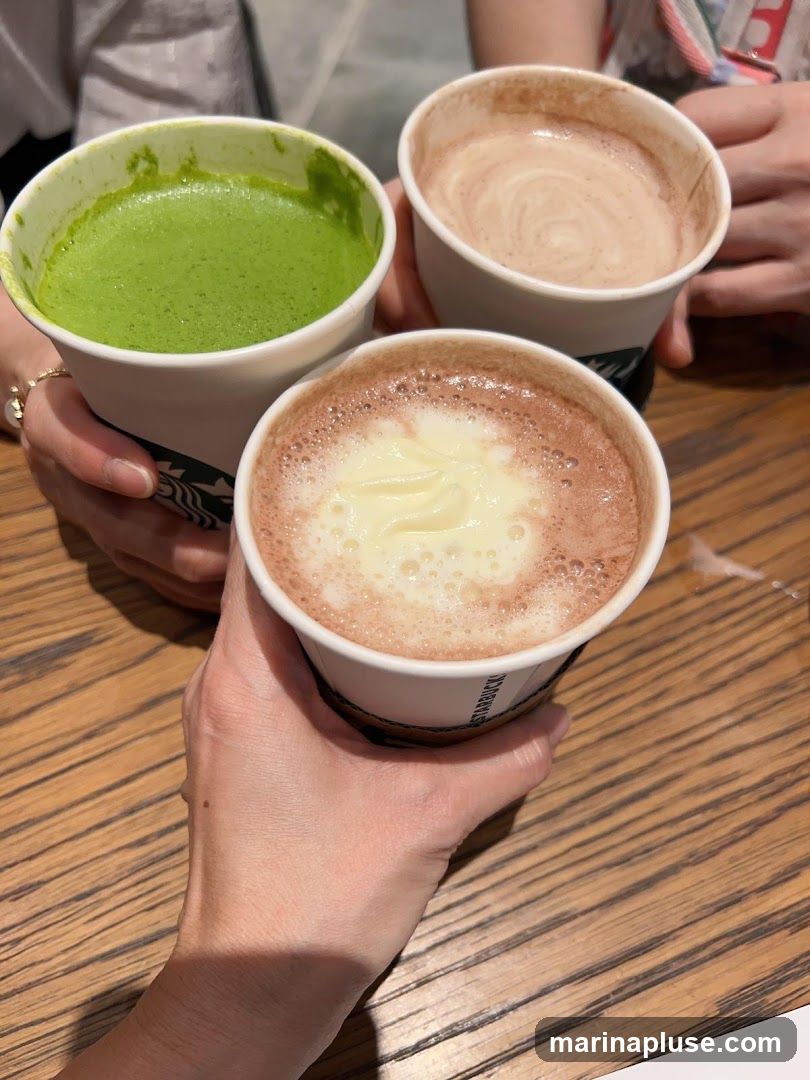 Three paper cups with drinks are held by hands. One cup contains a bright green beverage, likely matcha. Another cup has a light brown, swirled beverage. The third cup contains a darker brown beverage topped with white cream. The cups have the Starbucks logo visible.