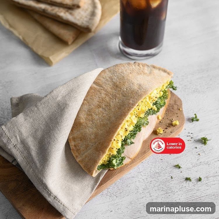 A sandwich filled with scrambled eggs and kale, served on a wooden board with a side of pita bread and a glass of iced coffee. A red circular sticker indicates "Lower in calories".