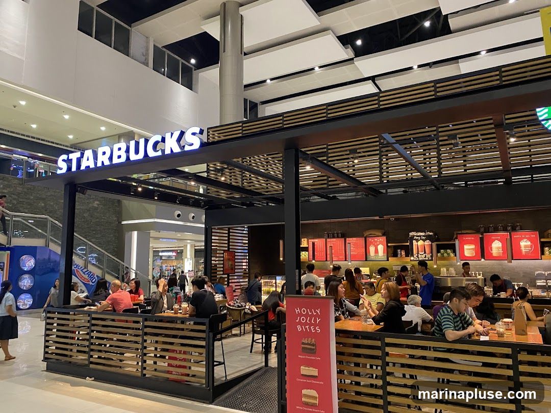 A busy Starbucks cafe with customers seated at tables both inside and on an outdoor patio area. The prominent Starbucks logo is visible above the entrance. The cafe features a modern design with wooden accents and warm lighting. A menu board displays various food and drink options, and promotional posters are visible on the walls.