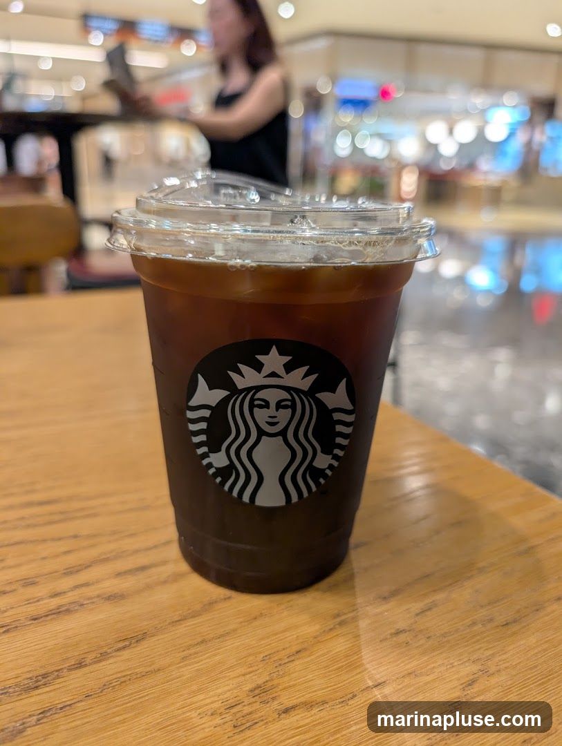 A close-up shot of an iced coffee in a clear plastic cup with a lid. The cup has the Starbucks logo on it. The drink is dark brown and filled with ice. The cup is sitting on a wooden table. In the blurred background, a person is visible, and the setting appears to be an indoor public space like a mall or cafe.