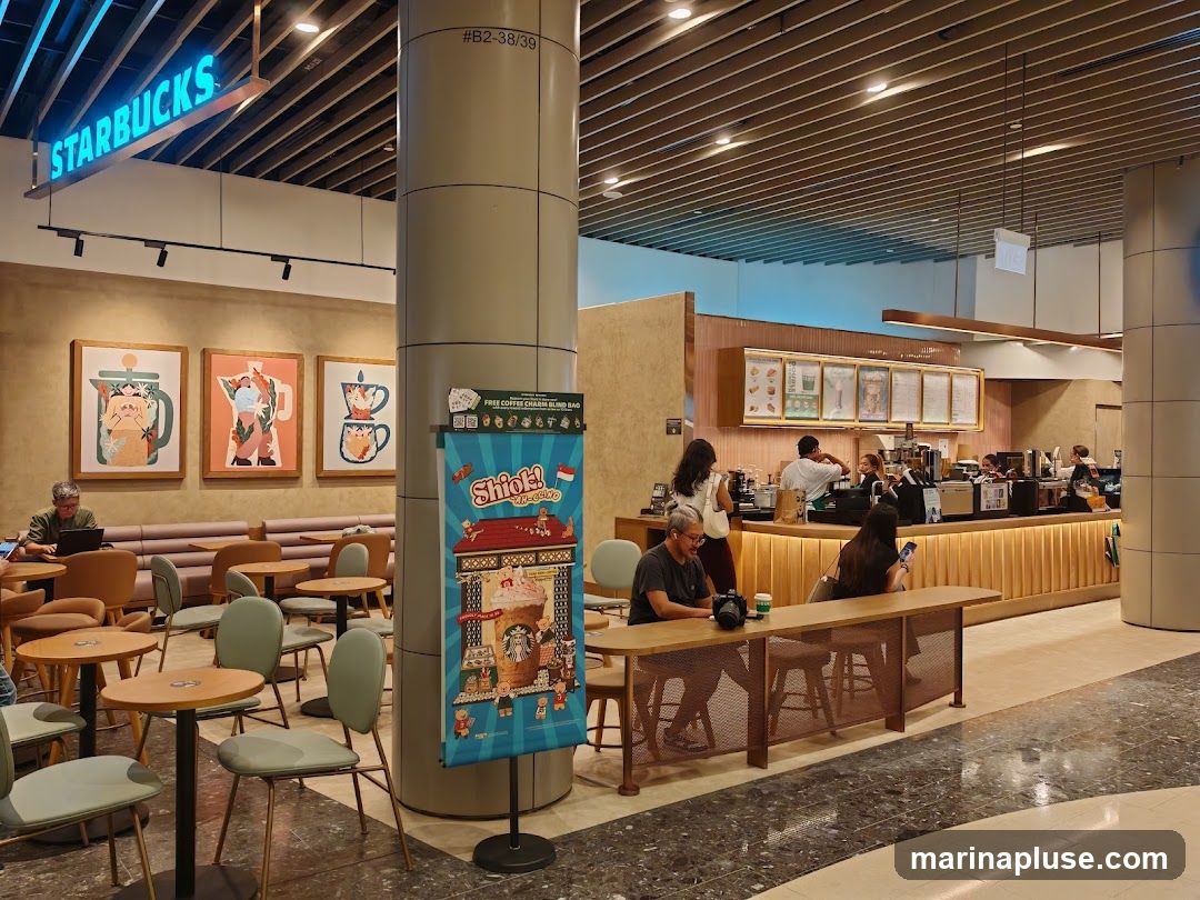 The interior of a Starbucks cafe with seating areas and a counter. A prominent "STARBUCKS" sign is visible on the ceiling. There are framed artworks on the wall and a promotional poster for a "Free Coffee Charm Blind Bag" with the text "Shiok!". Customers are seated at tables and at the counter.