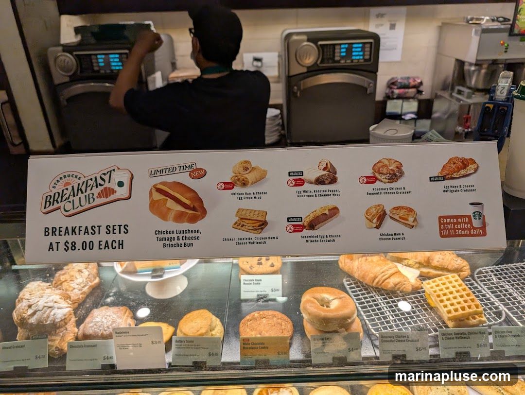 A sign displays Starbucks Breakfast Club sets for $8.00 each, featuring various sandwiches and wraps. Below the sign, a display case shows a selection of pastries and breakfast items.