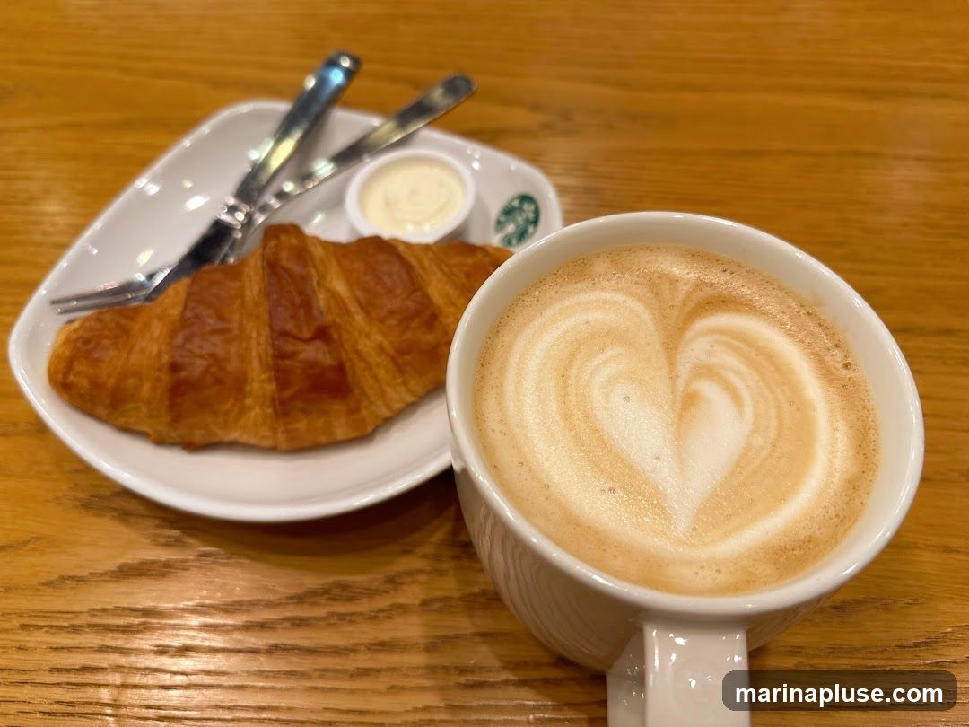 A golden-brown croissant is served on a white plate with a small container of cream and silverware. Next to the plate is a white mug filled with a latte, featuring a heart design in the foam. The food and drink are placed on a wooden table. A faint Starbucks logo is visible on the plate.