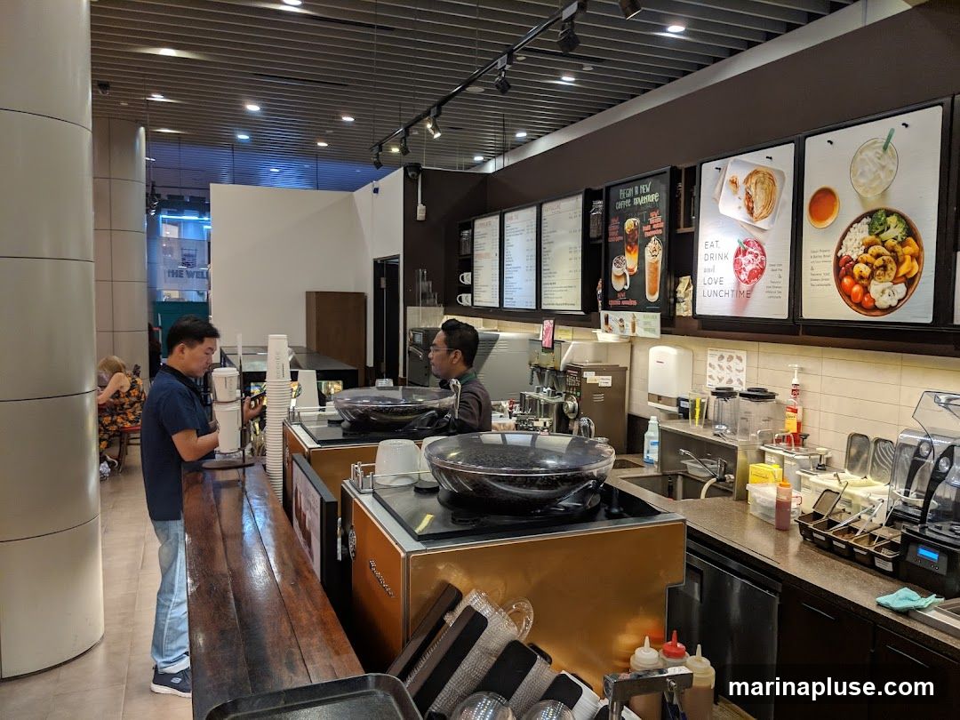 A view of a coffee shop counter with baristas preparing drinks and customers ordering. There are menu boards displayed above the counter, showing various food and drink options. The counter is equipped with coffee machines, grinders, and other necessary equipment.