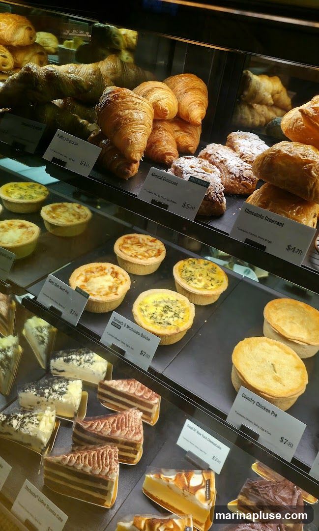 A display case filled with various pastries and cakes. Items include croissants, quiches, pies, and layered cakes. Labels indicate prices and some ingredients.