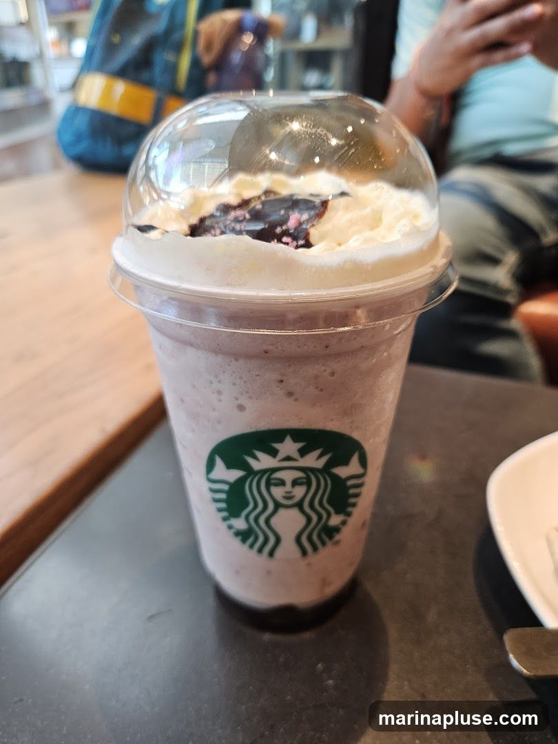 A close-up of a Starbucks Frappuccino with whipped cream and a dark topping, featuring the iconic Starbucks logo on the cup. The drink is on a dark surface, with a wooden table and blurred figures in the background.