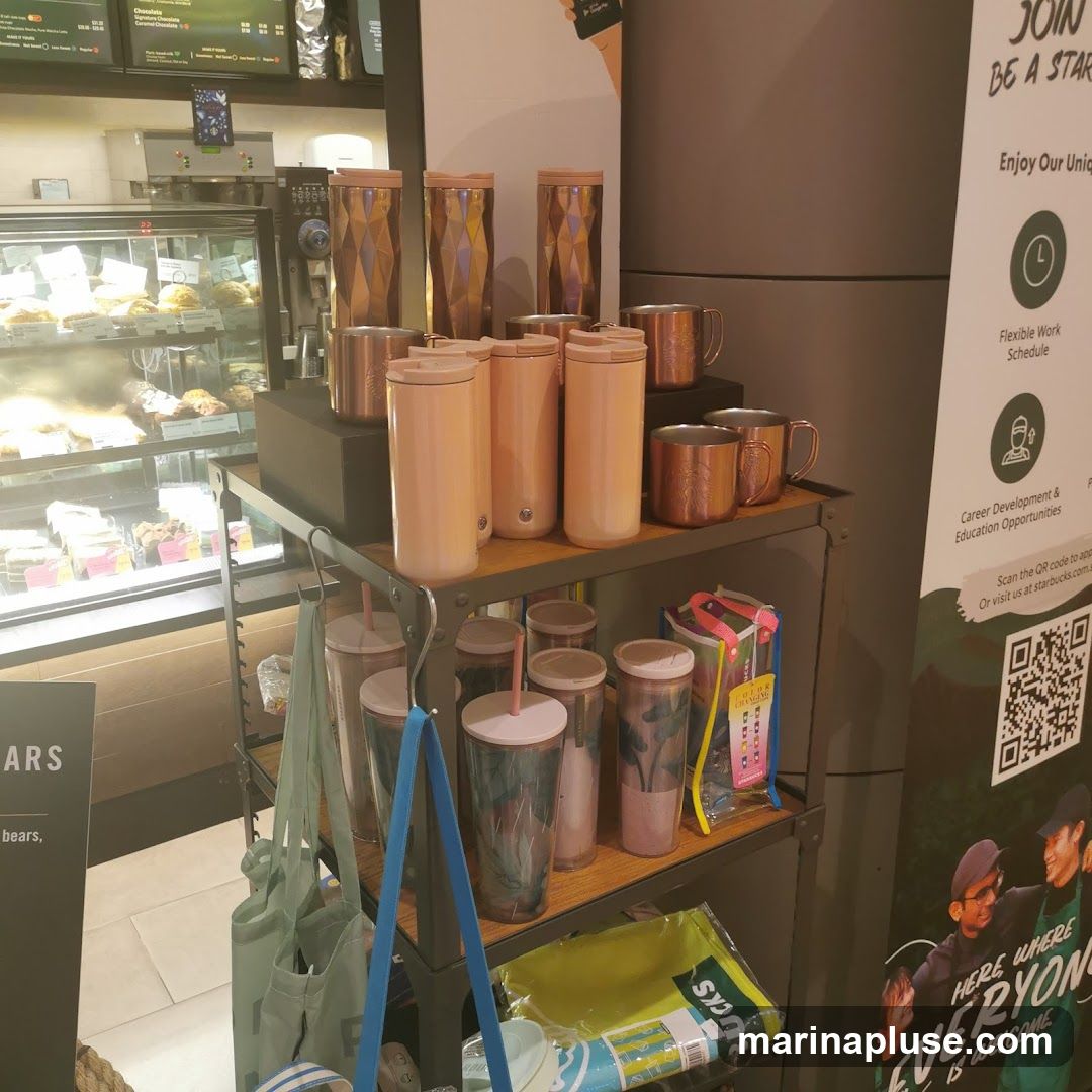 A display of Starbucks merchandise including tumblers, mugs, and reusable bags. In the background, a refrigerated display case shows baked goods, and a menu board is visible above. A recruitment poster for Starbucks is on the right side of the image.