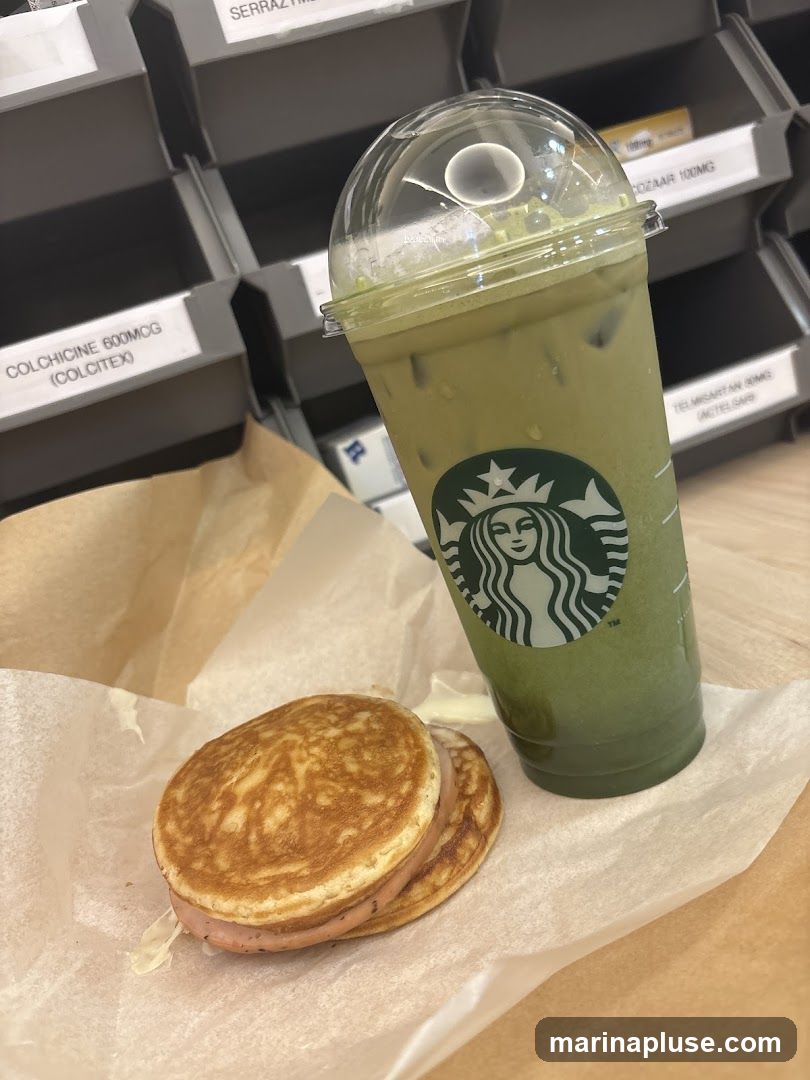 A Starbucks iced matcha latte with whipped cream and a pancake sandwich with ham filling are displayed on a counter. In the background, shelves are filled with various pharmaceutical products.