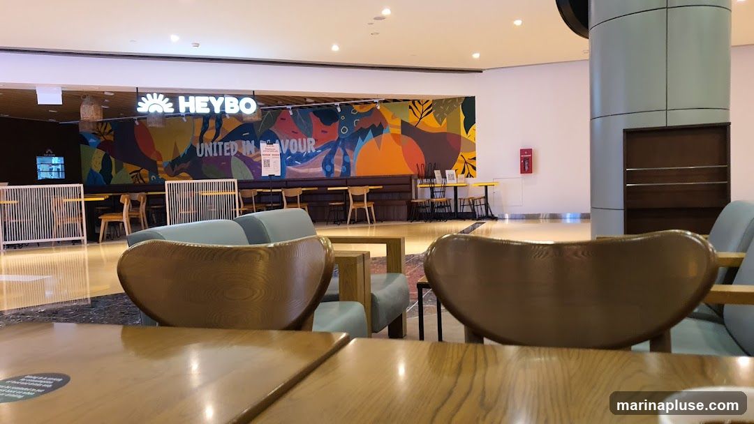 The image shows the interior of a cafe with a prominent, colorful mural on the back wall. The mural features abstract shapes and natural elements in vibrant colors like orange, yellow, blue, and green. Above the mural, the cafe's name, "HEYBO," is displayed in illuminated white lettering, accompanied by a sun-like logo. The seating area includes light-colored wooden chairs and tables, some with white dividers, and comfortable-looking light blue sofas. The floor is highly reflective, mirroring the lights and furniture. In the background, there are more tables and chairs, and a large cylindrical pillar is visible on the right side.
