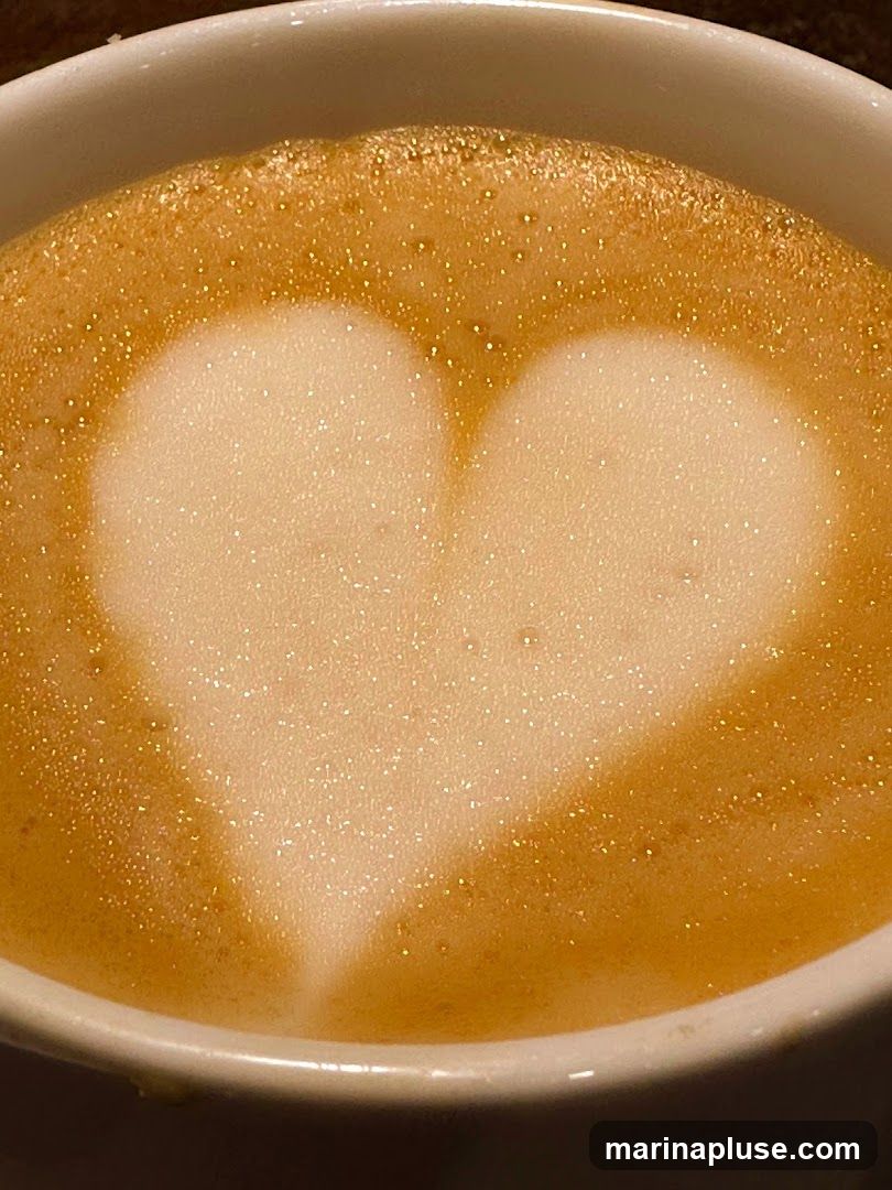 A close-up shot of a white mug filled with a latte. The surface of the latte has a heart shape created with foam art. The foam is light and airy, with small bubbles visible. The coffee beneath the foam is a rich brown color.