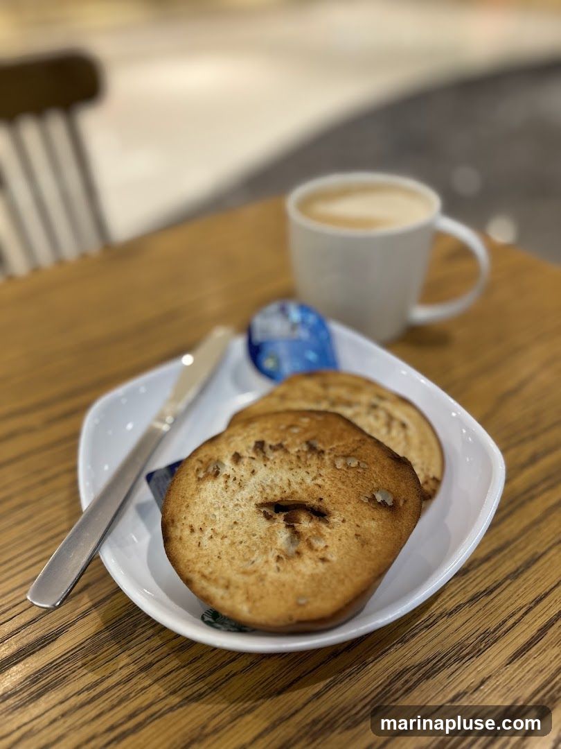 A white plate holds two toasted bagel halves, a small packet of butter, and a knife. A white mug of coffee with latte art sits in the background on a wooden table.