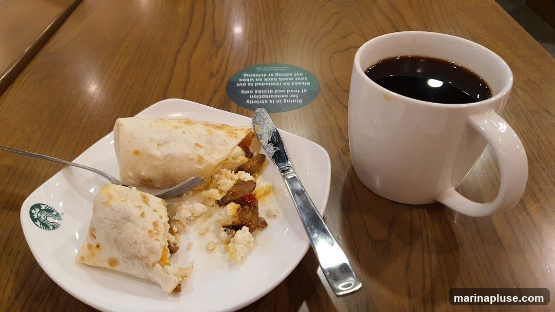A white plate with a partially eaten burrito and a fork and knife sits on a wooden table. A white mug filled with black coffee is to the right of the plate. A green and white circular sticker on the table reads: "Dining in is strictly for consumption of food and drinks only. Please be reminded to put your mask back on when not eating or drinking." The Starbucks logo is visible on the plate.