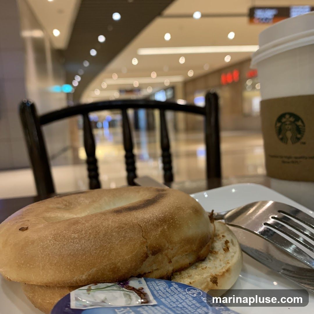 A close-up shot of a bagel and a cup of coffee on a white plate, with a fork resting on the side. The coffee cup has a Starbucks logo on its sleeve. The background shows a blurred indoor environment, possibly a mall or food court, with bright lighting and seating areas.