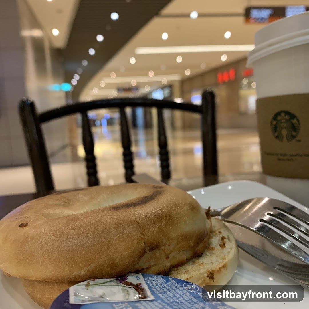 A close-up shot of a bagel and a cup of coffee on a white plate, with a fork resting on the side. The coffee cup has a Starbucks logo on its sleeve. The background shows a blurred indoor environment, possibly a mall or food court, with bright lighting and seating areas.