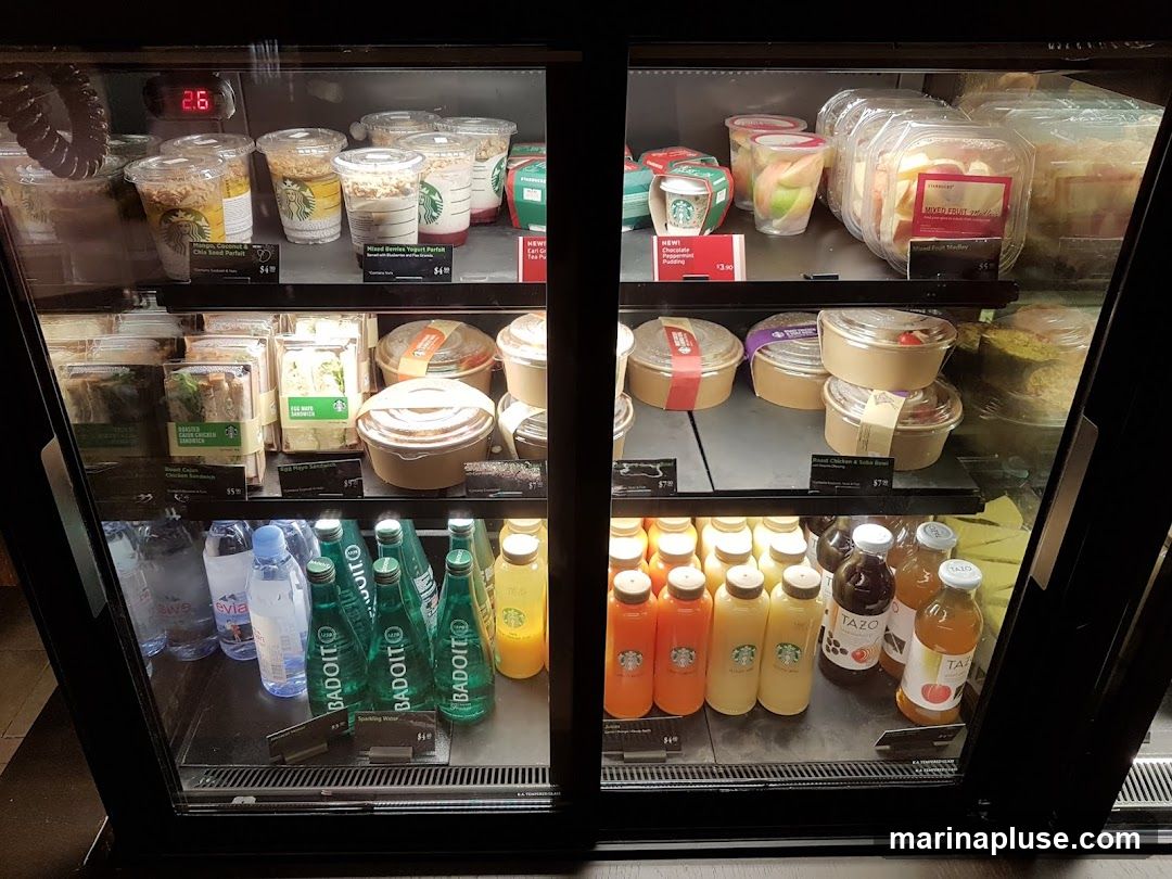 A refrigerated display case filled with various food and beverage items. The top shelf contains yogurt parfaits and fruit cups. The middle shelves hold sandwiches, salads, and bowls. The bottom shelf displays bottled water, sparkling water, juices, and iced tea.