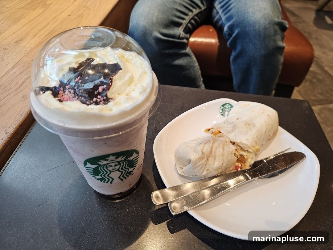 A close-up shot shows a Starbucks Frappuccino topped with whipped cream, chocolate drizzle, and pink sprinkles. Next to it, on a white plate with the Starbucks logo, is a wrap that has been cut in half, revealing its filling. A fork and knife are placed on the plate. In the background, a person wearing blue jeans is seated on a brown chair.