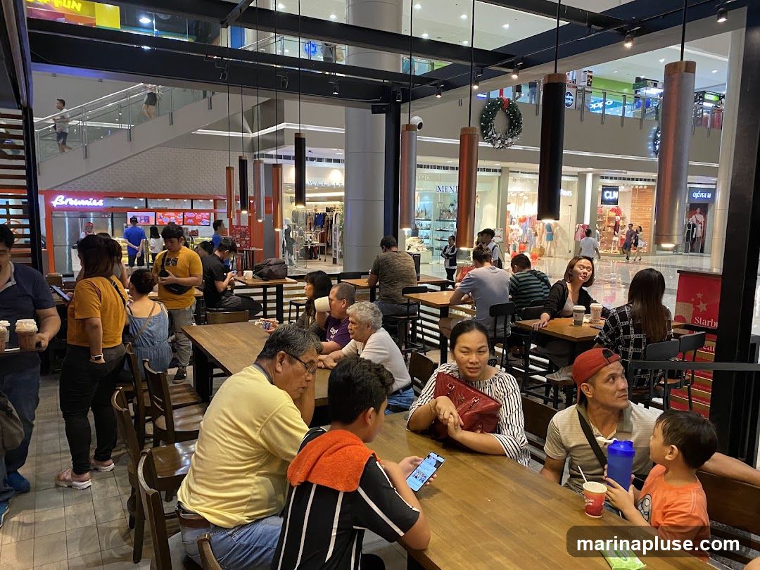 A group of people are seated at wooden tables in a cafe, with some looking at their phones and others conversing. In the background, there are shops and escalators within a mall.