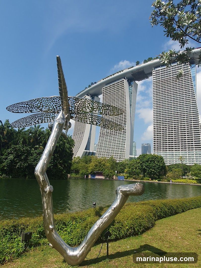 A large, metallic sculpture of a dragonfly is positioned in the foreground of a park. The sculpture is abstract, with intricate patterns on its wings. Behind the sculpture, a calm body of water reflects the sky and surrounding greenery. In the distance, the iconic Marina Bay Sands hotel complex stands tall, its distinctive architecture dominating the skyline under a clear blue sky with scattered clouds. Lush trees and manicured bushes complete the scenic view.