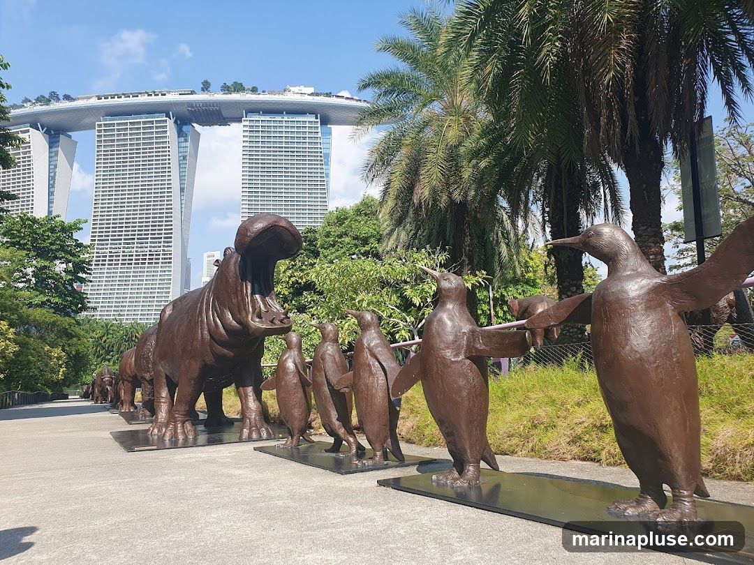 A line of bronze animal sculptures, including a hippopotamus and penguins, are displayed outdoors on a paved walkway. In the background, the distinctive Marina Bay Sands hotel complex is visible, along with palm trees and lush greenery under a clear blue sky.