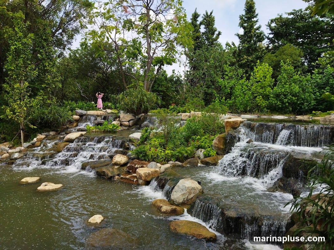 A serene landscape featuring a multi-tiered waterfall cascading over rocks into a pool of water. Lush green trees and foliage surround the water feature, creating a tranquil natural setting. A woman in a pink dress stands on a rock formation near the top of the waterfall, appearing to admire the view.