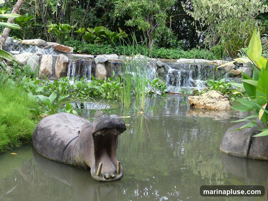 A concrete hippopotamus sculpture with its mouth open sits in a pond with a waterfall in the background. Lush greenery surrounds the water feature.