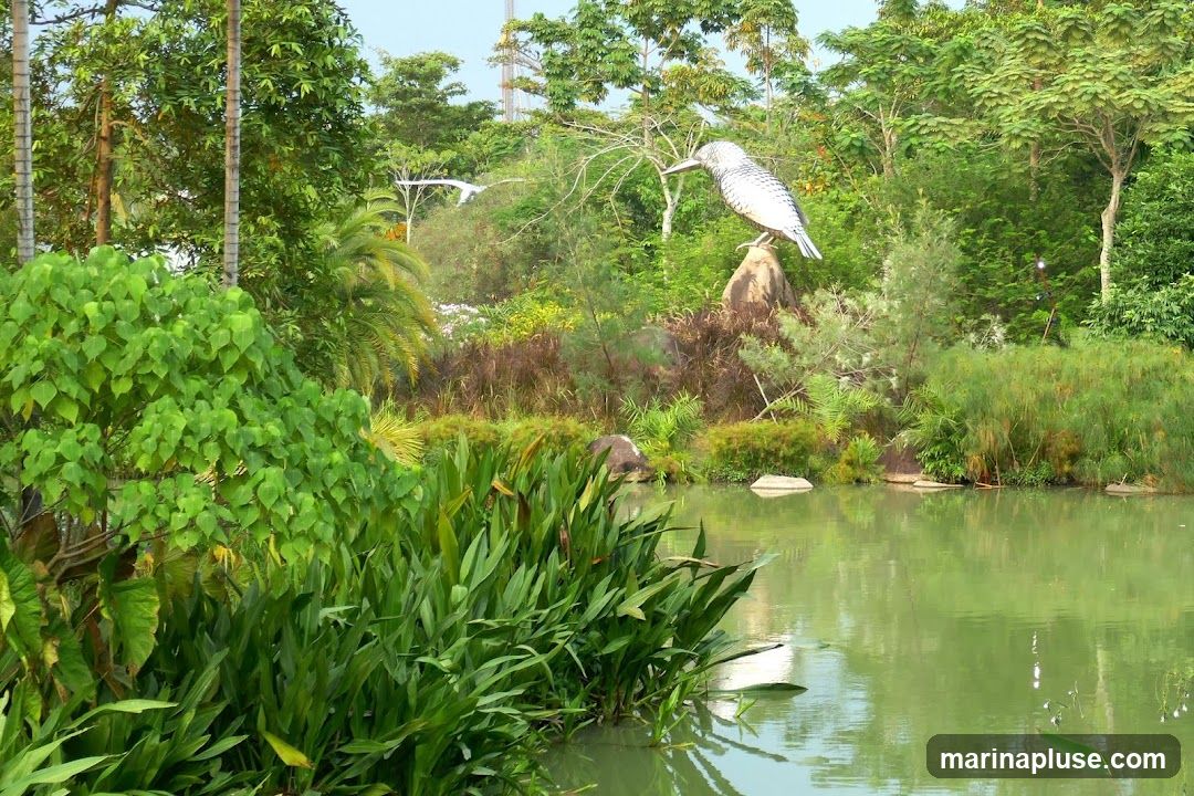 A large, metallic sculpture of a bird, possibly a kingfisher, stands on a rock overlooking a calm body of water. The sculpture is detailed with a patterned body and a long beak. The surrounding environment is lush and green, with various types of trees, plants, and reeds growing along the water's edge. In the background, two white bird sculptures are in flight, and a tall tower-like structure is visible above the trees. The water reflects the greenery and the sky, creating a serene natural scene with artistic elements.
