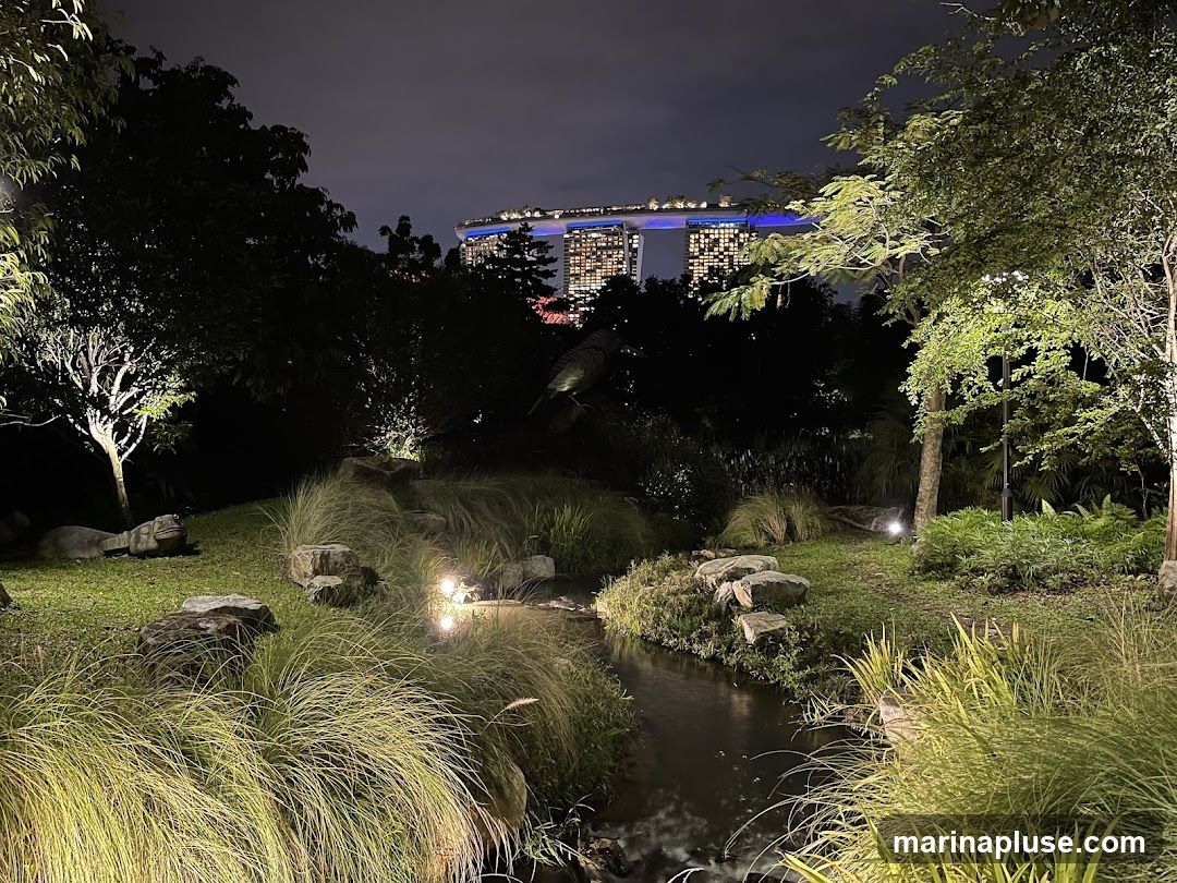 A serene garden scene at night, with lush greenery, a small stream, and rocks illuminated by soft lighting. In the background, the illuminated skyline of a city, featuring distinctive skyscrapers, is visible against the dark sky.