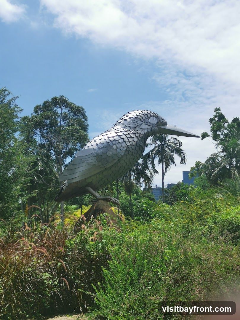 A large, metallic sculpture of a kingfisher bird stands prominently in a vibrant green garden. The sculpture is detailed, with individual metallic scales forming its body and wings. It is perched on a rock or log, surrounded by lush foliage, including tall grasses, bushes, and palm trees. In the background, a modern cityscape is visible through the trees under a partly cloudy blue sky.