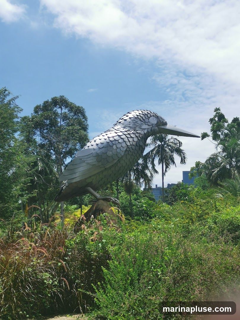 A large, metallic sculpture of a kingfisher bird stands prominently in a vibrant green garden. The sculpture is detailed, with individual metallic scales forming its body and wings. It is perched on a rock or log, surrounded by lush foliage, including tall grasses, bushes, and palm trees. In the background, a modern cityscape is visible through the trees under a partly cloudy blue sky.