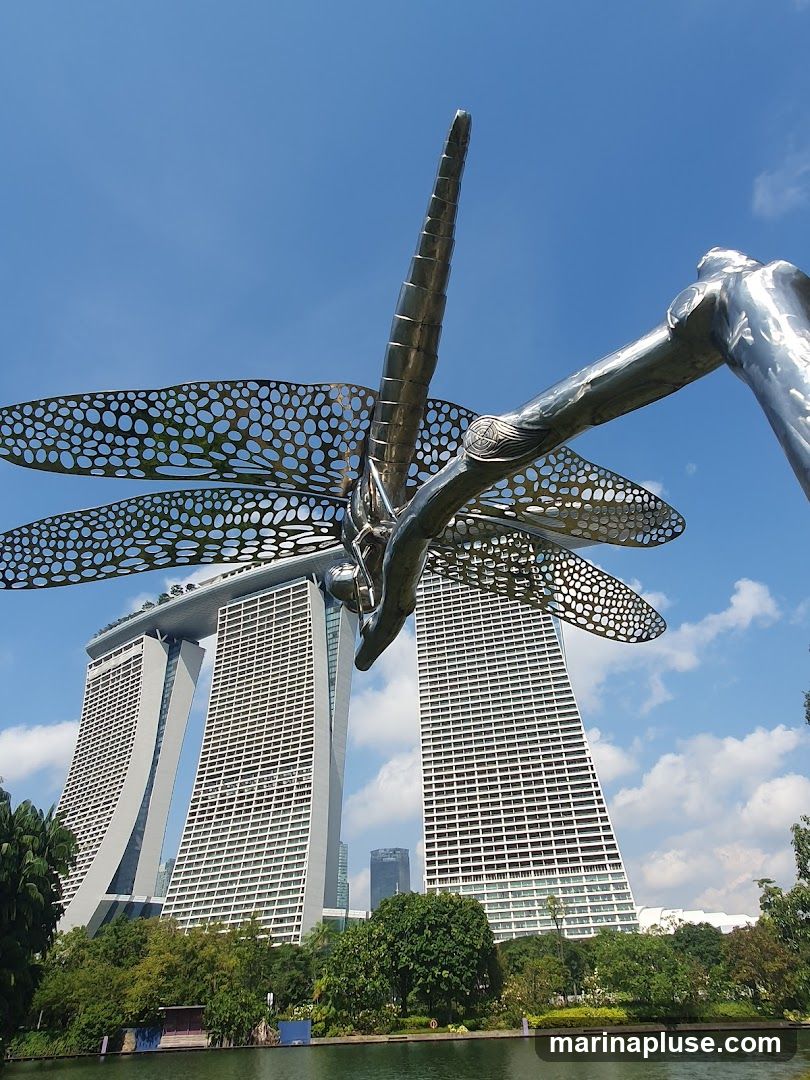 A large, metallic sculpture of a dragonfly is prominently featured in the foreground, with its intricate wings spread wide. In the background, the iconic Marina Bay Sands hotel complex stands tall against a clear blue sky with scattered clouds. Lush green trees and a body of water are visible below the buildings, suggesting a park or garden setting.