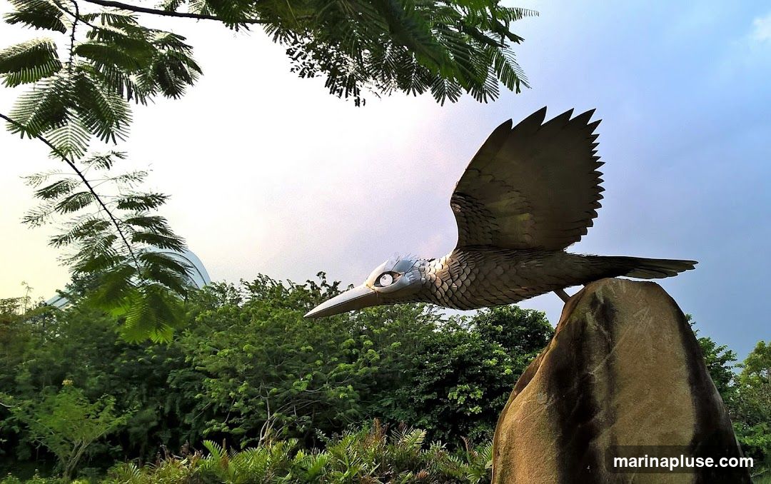 A metallic sculpture of a bird-like dinosaur in flight is perched on a rock. The sculpture is detailed with scales and has large, reflective eyes. It is set against a backdrop of lush green trees and a cloudy sky. In the distance, a large, curved structure, possibly a dome or greenhouse, is visible through the foliage.