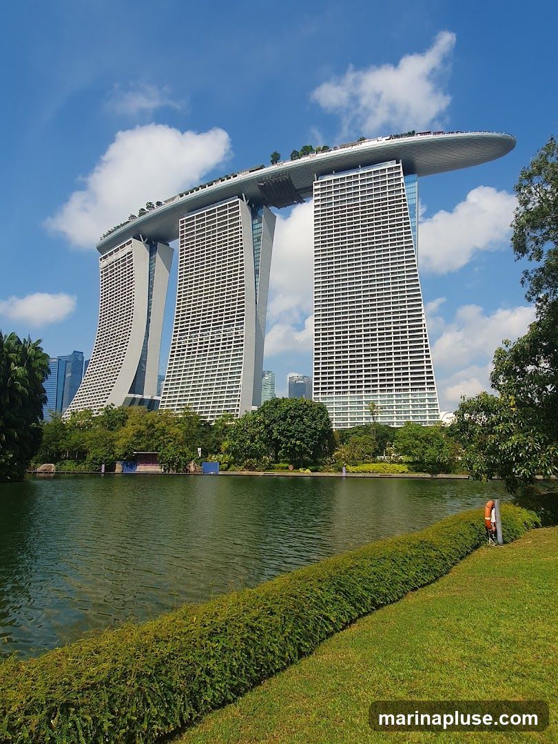 A wide shot of the Marina Bay Sands hotel in Singapore, with its distinctive three towers topped by a large, boat-shaped deck. The hotel is set against a bright blue sky with scattered white clouds. In the foreground, a calm body of water reflects the sky and the surrounding greenery. A neatly trimmed hedge runs along the edge of the water, separating it from a lush green lawn. Trees and other buildings are visible in the background.