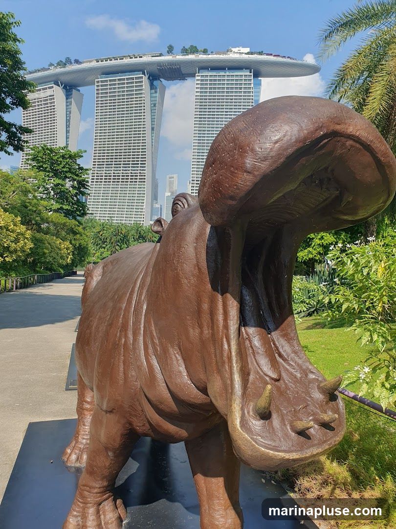 A bronze statue of a hippopotamus with its mouth open stands in a park. In the background, the iconic Marina Bay Sands hotel complex is visible under a clear blue sky. Lush greenery and palm trees surround the statue and the buildings.