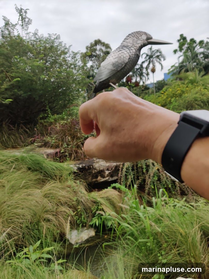 A close-up shot of a person's hand in the foreground, with a metallic sculpture of a bird perched on their finger. The background features lush greenery, including tall grasses, bushes, trees, and a small stream. The sky is overcast.