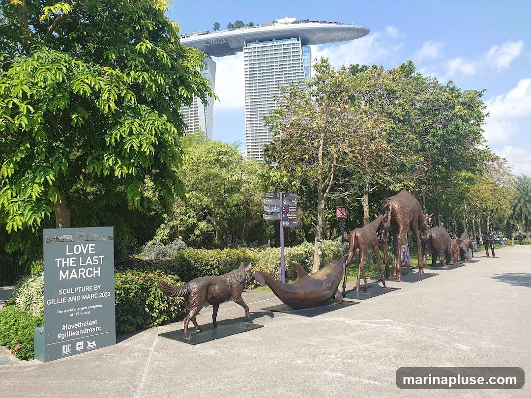 A bronze sculpture of various animals, including a dolphin, wolf, and giraffes, is displayed outdoors. A sign in the foreground reads "LOVE THE LAST MARCH SCULPTURE BY GILLIE AND MARC 2023 The world's largest sculpture at 192m long #lovethelast #gillieandmarc". In the background, the Marina Bay Sands hotel is visible.