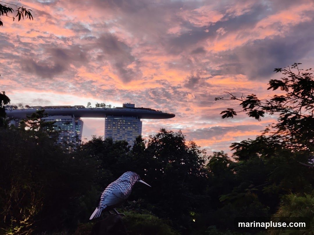 A scenic view of the Marina Bay Sands hotel in Singapore during sunset. The sky is filled with dramatic clouds painted in shades of pink, orange, and purple. In the foreground, silhouetted trees and a decorative bird sculpture add depth to the scene.
