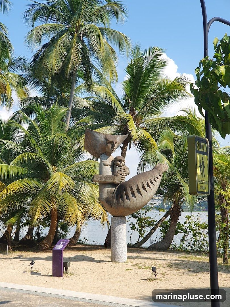 A stone sculpture of a figure holding a child is displayed outdoors, surrounded by palm trees and a sandy ground. A body of water is visible in the background, along with a pedestrian crossing sign.