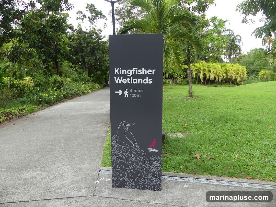 A dark gray sign with white text and graphics stands on a paved path in a park. The sign reads "Kingfisher Wetlands" with an arrow pointing right, indicating "4 mins" and "120m". Below this, there is a line drawing of a bird and floral patterns. At the bottom of the sign is the logo for "Gardens by the Bay". The background shows a lush green lawn, trees, and a pathway curving to the left.