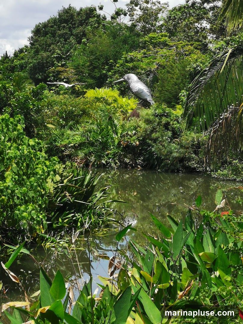 A scenic view of a garden with a body of water in the foreground. Lush green foliage surrounds the water, with tall trees forming a dense canopy in the background. Two bird sculptures are visible amidst the greenery; one is a large, metallic sculpture of a bird with its wings spread, perched on a mound. Another, smaller white bird sculpture appears to be in flight. The water reflects the surrounding vegetation and the sky.