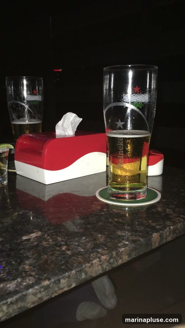 Two glasses of beer sit on a granite bar top. A red and white tissue box is between them. One glass is nearly full, the other is half empty. A shot glass with a lime wedge is visible to the left. The background is dark.