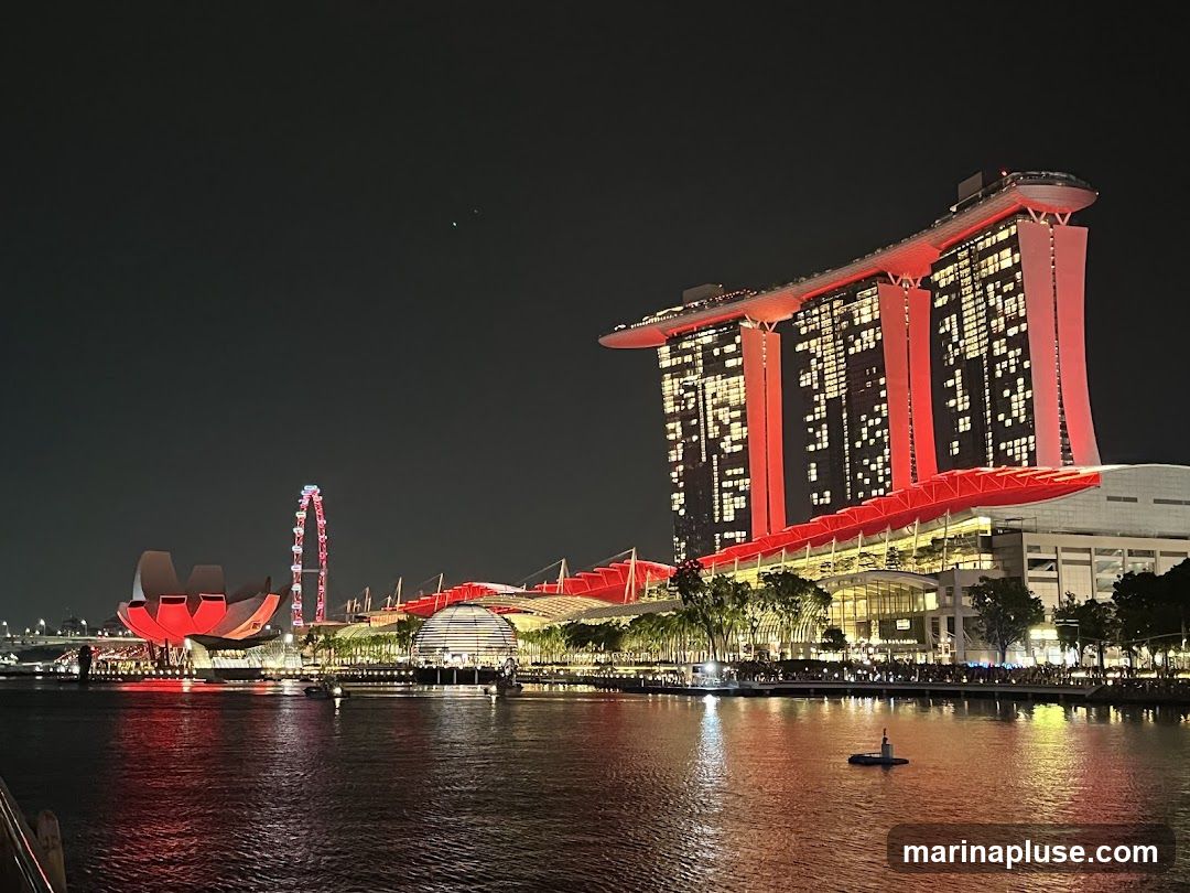 A nighttime view of the Marina Bay Sands complex in Singapore, illuminated with red lights. The iconic three towers are visible, connected by the SkyPark. In the foreground, a body of water reflects the lights. To the left, the ArtScience Museum and the Singapore Flyer Ferris wheel are also illuminated. The scene depicts a vibrant urban landscape at night.