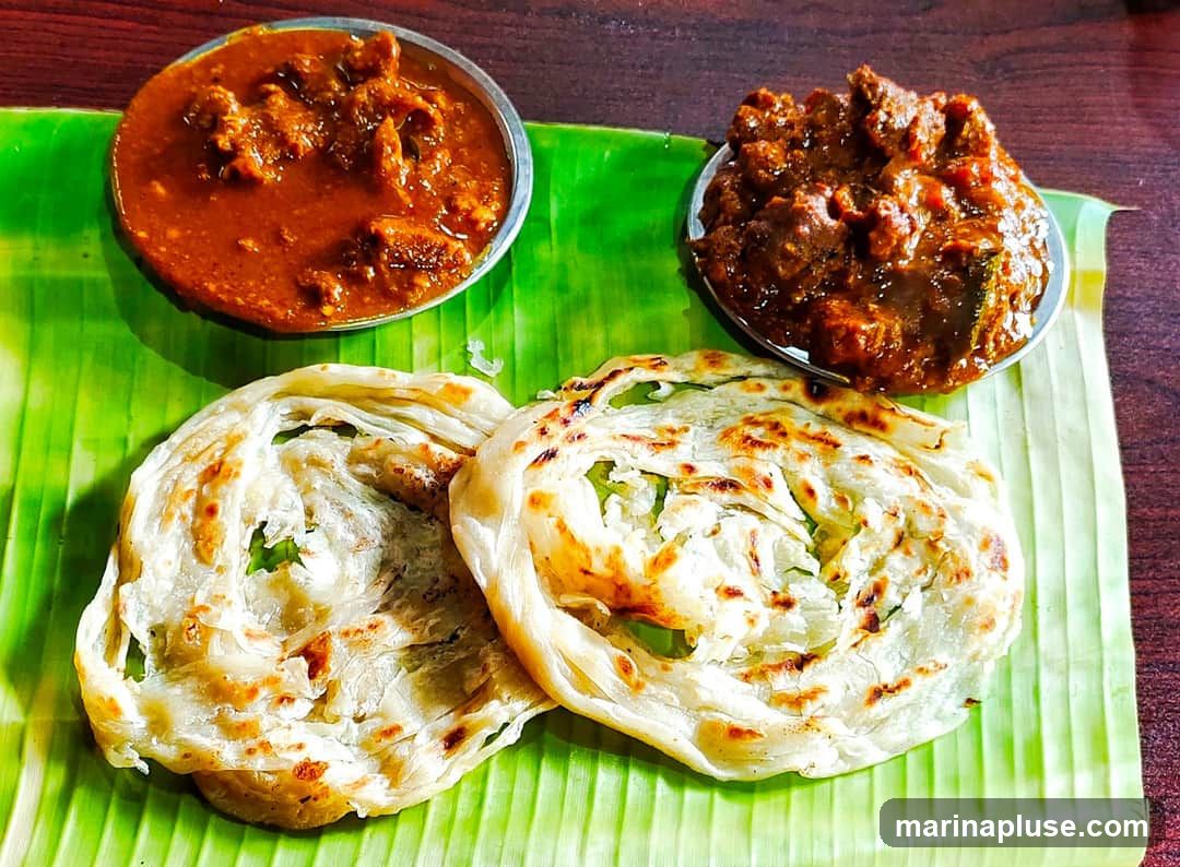 Two servings of parotta, a layered flatbread, are presented on a banana leaf alongside two bowls of curry. One curry is a rich, reddish-brown, while the other is a darker, more intense brown. The parottas are golden brown with visible layers and some charring.