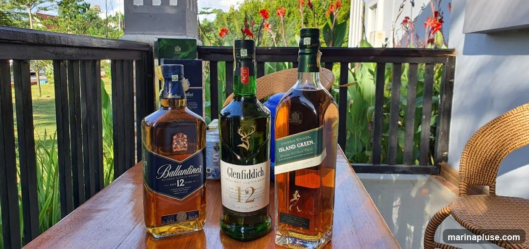 Three bottles of whiskey are displayed on a wooden table outdoors. From left to right, the bottles are Ballantine's 12 Years, Glenfiddich 12 Years, and Johnnie Walker Island Green. In the background, there is a railing, lush greenery, and red flowers. A wicker chair is visible on the right side of the image.