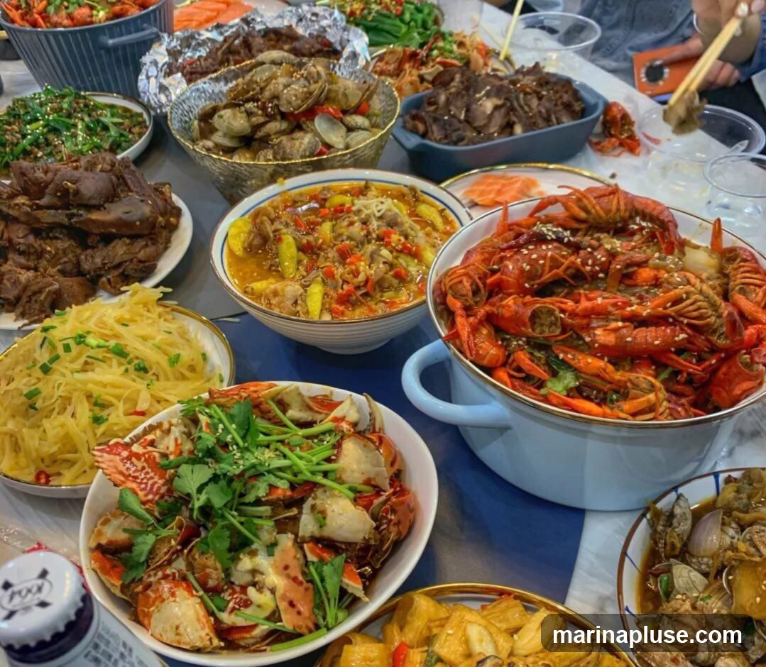 A table laden with various Chinese dishes, including a large bowl of crawfish, a platter of crab, stir-fried vegetables, shredded potatoes, clams, and sliced meat. The food is arranged in bowls and plates, suggesting a communal dining experience.